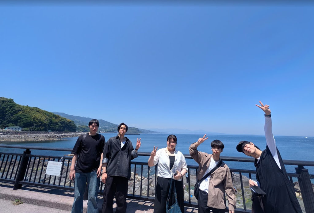 avinton-engineers-manazuru-ocean-drive 神奈川県真鶴町の広大な青い海と空をバックに、ピースサインをしてリフレッシュするAvintonの若手エンジニアたちの集合写真。