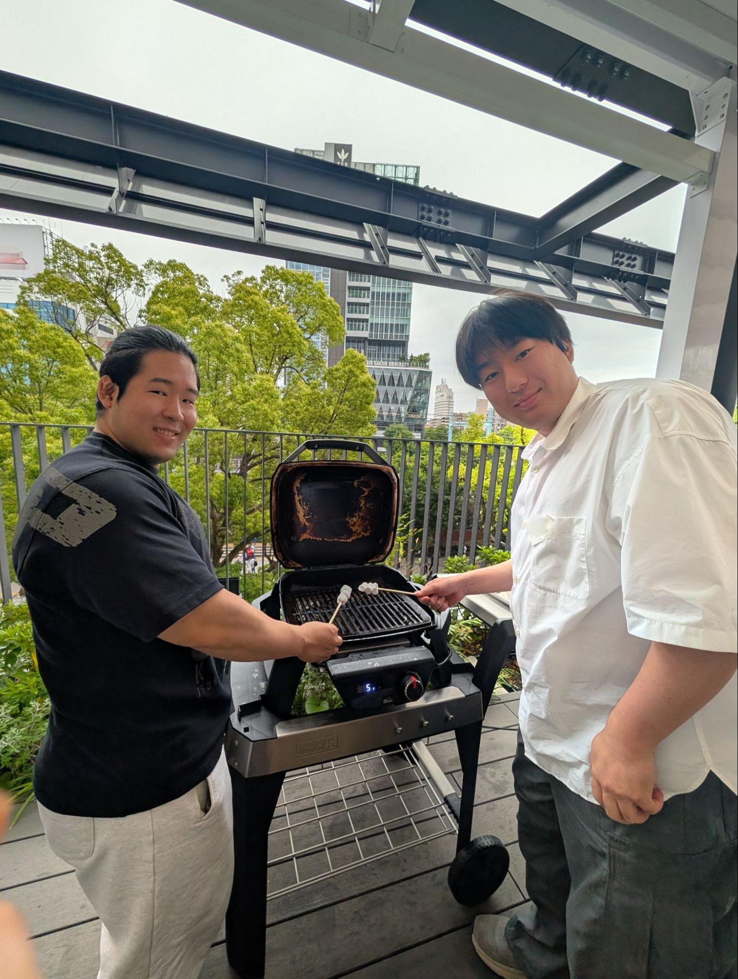 会社のBBQイベントで、テラスのグリルを囲んで楽しそうにマシュマロを焼く社員たちの様子